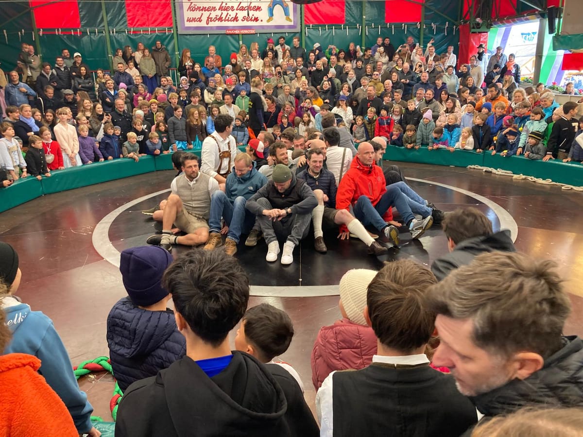 A crowd packed around the rim of the Teufelsrad, the spinning wooden disc ride at Munich's Oktoberfest, watching a cluster of grown men sitting huddled together at its center