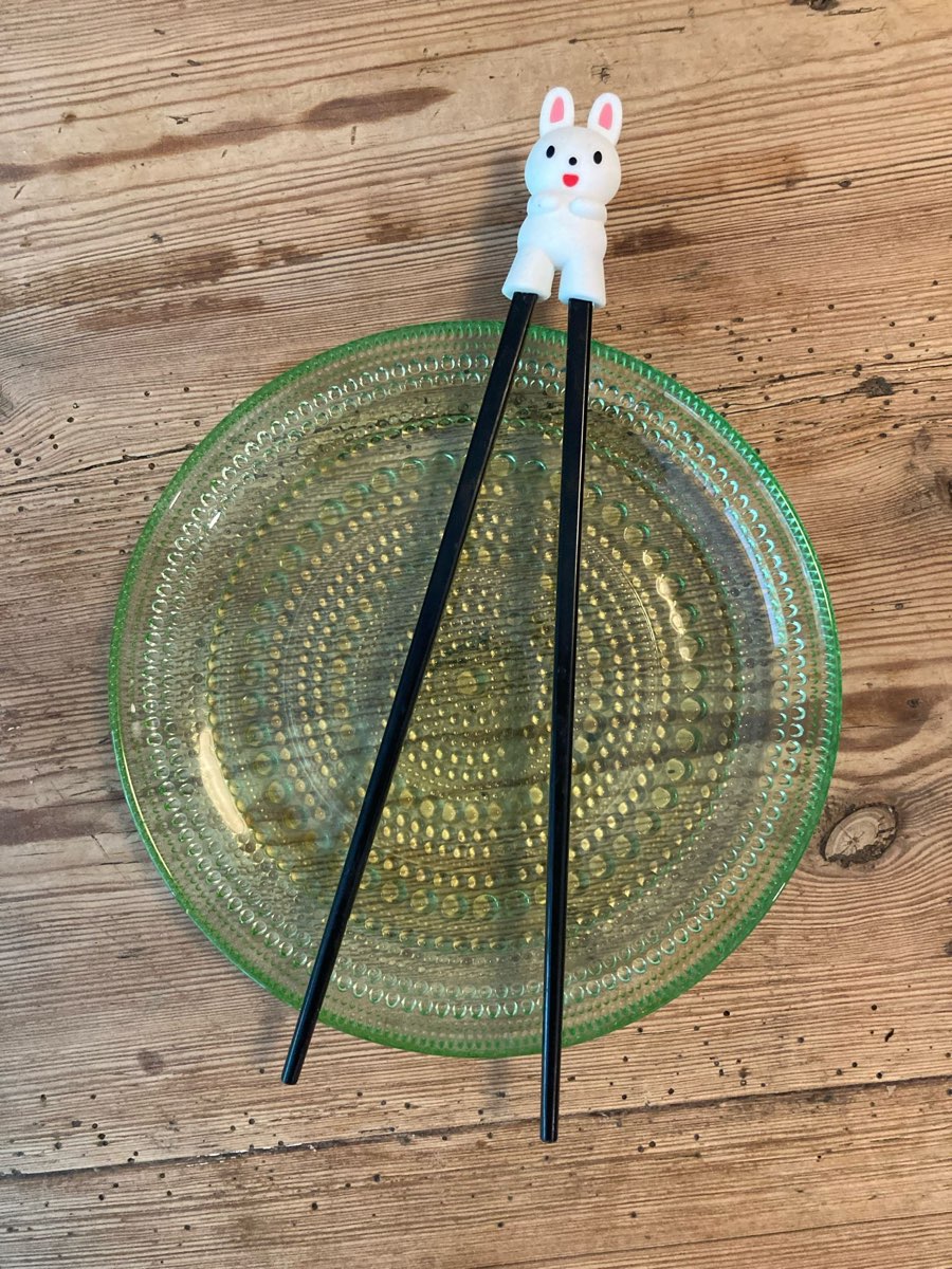 A bunny-topped children's chopstick helper resting on a green glass plate on a wooden table