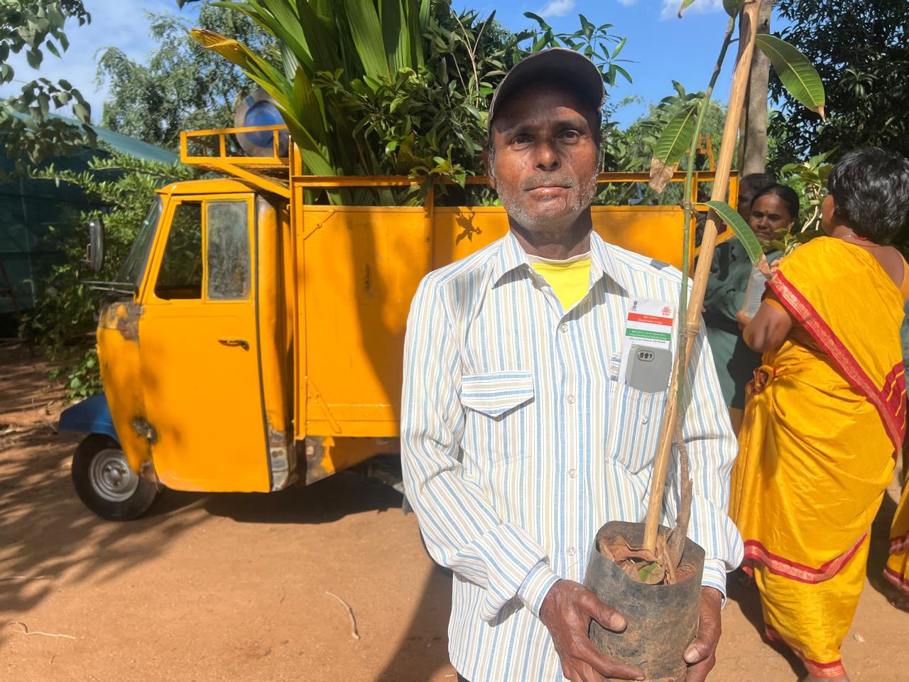 A farmer in a cap holding a potted fruit-tree sapling in front of a yellow auto rickshaw loaded with saplings