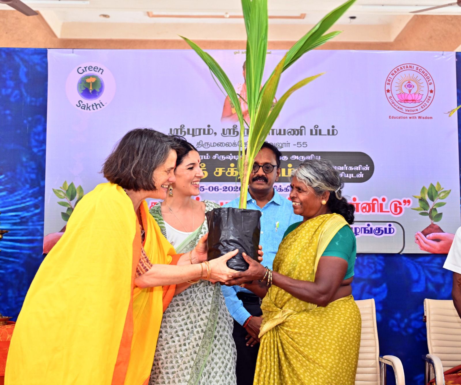Nathalie Latham and another woman handing a potted coconut sapling to a farmer at a Green Sakthi giveaway, an event banner behind them